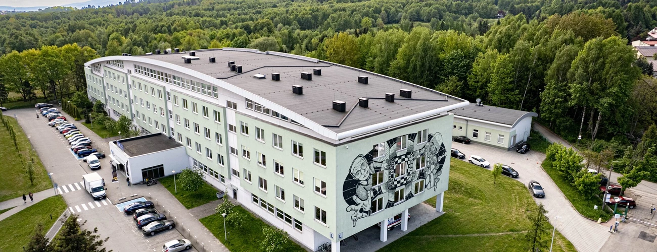 Widok z lotu ptaka na wielopiętrowy budynek „L” Uniwersytetu Bielsko-Bialskiego. Budynek otoczony jest zielenią, z licznymi zaparkowanymi samochodami przy wejściu. W tle widać góry porośnięte drzewami.